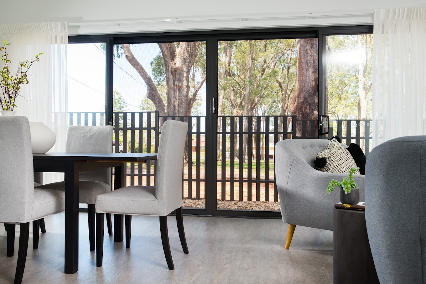 Bright living and dining area with large black-framed sliding glass doors opening to a balcony, with trees visible outside.